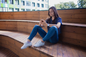 A brunette girl, a student with a large wrist watch, sits on wooden steps with legs and thoughtfully checks smartphone.